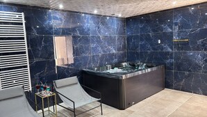 A modern bathroom with a marble wall, a black jacuzzi, a glass-walled shower, and a small table with toiletries.