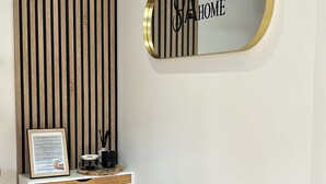 A modern reception area with a vertical wooden panel wall, a reception desk with a framed document, a black vase with flowers, and a mirror with 'SA HOME' on it.