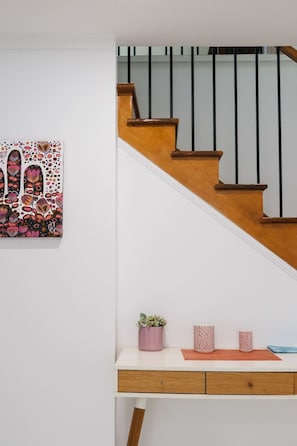 Een trap met houten treden en zwarte balustrades, een witte muur met een kleurrijk abstract schilderij, en een klein houten bureau met een plant en decoratieve potten.