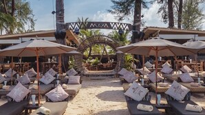 Un resort frente a la playa con tumbonas y sombrillas, rodeado de altos árboles y un cielo despejado.