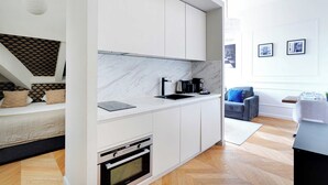 A modern kitchen with white cabinets, a built-in oven, and a marble backsplash. There is a dining area with a gray sofa and a small table. - Magnificent Apartment -1br-4p- Champs-elysees (Paris)