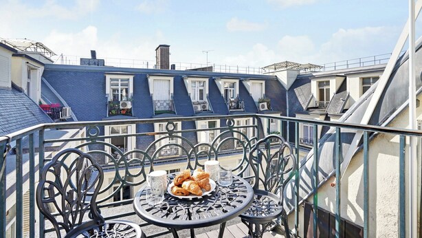 A balcony with a table set for breakfast, featuring croissants and glasses, overlooking a row of blue buildings with white windows. - Magnificent Apartment -1br-4p- Champs-elysees (Paris)