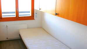 Une petite chambre avec un lit simple, une armoire en bois et une fenêtre donnant sur le ciel.
