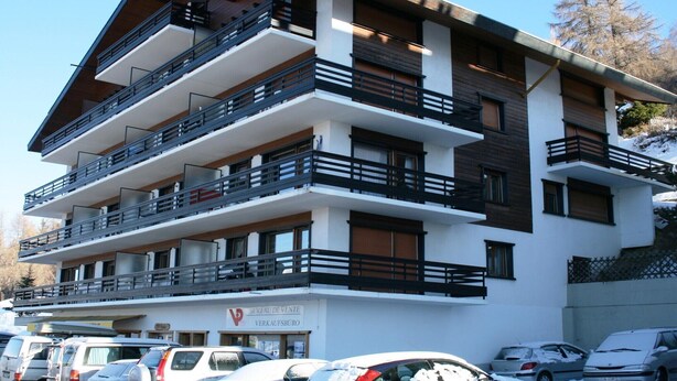 Un immeuble à plusieurs étages doté de balcons, une enseigne indiquant qu’il s’agit d’un bureau de vente, et des voitures garées devant.