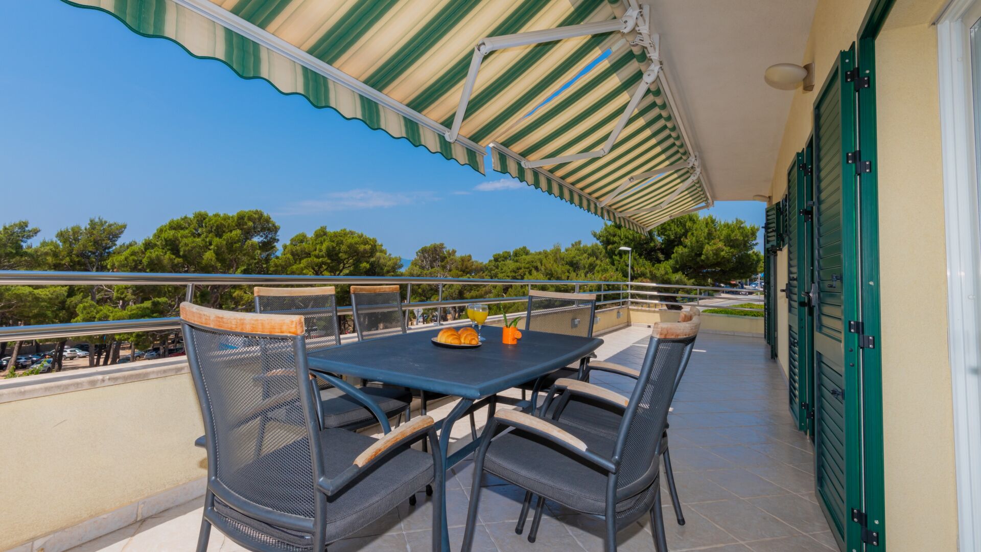 A balcony with a table and chairs, a striped awning, and a view of trees and a parking lot.