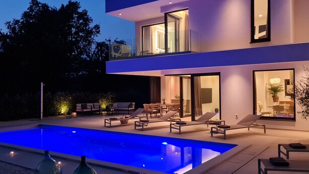 Una casa moderna de dos pisos amb piscina, mobiliari exterior i interior ben il·luminat.
