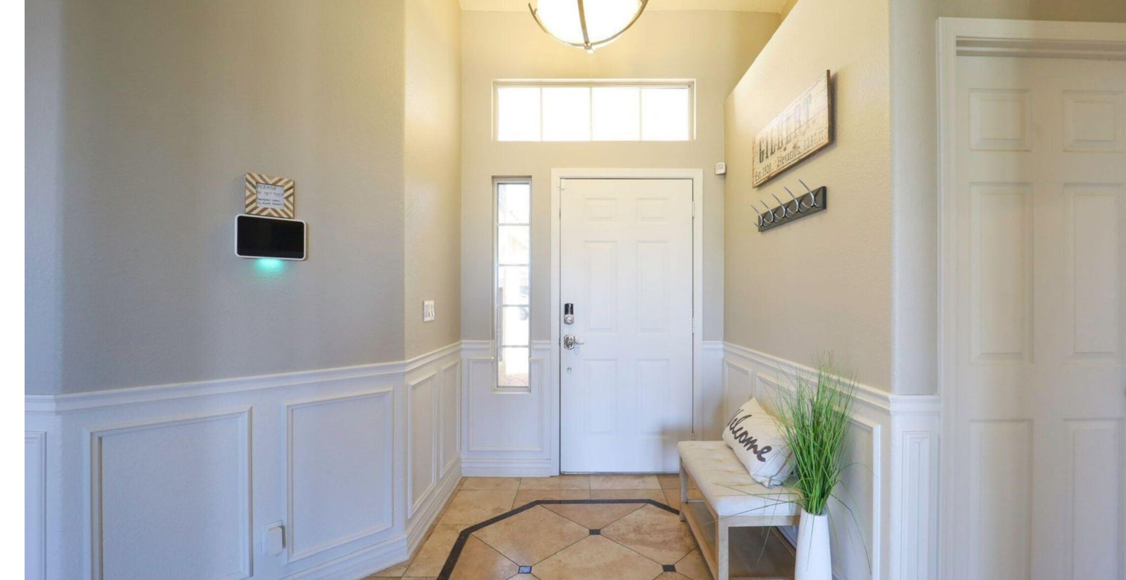 A hallway with a white door, a bench with a 'Welcome' pillow, a wall-mounted sign, and a light fixture.