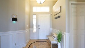 A hallway with a white door, a bench with a 'Welcome' pillow, a wall-mounted sign, and a light fixture.