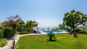 A pool area with a view of the sea, surrounded by greenery and a red umbrella.