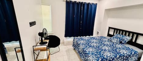Un dormitorio con cama, escritorio, silla, ventilador y ventana con cortinas.
