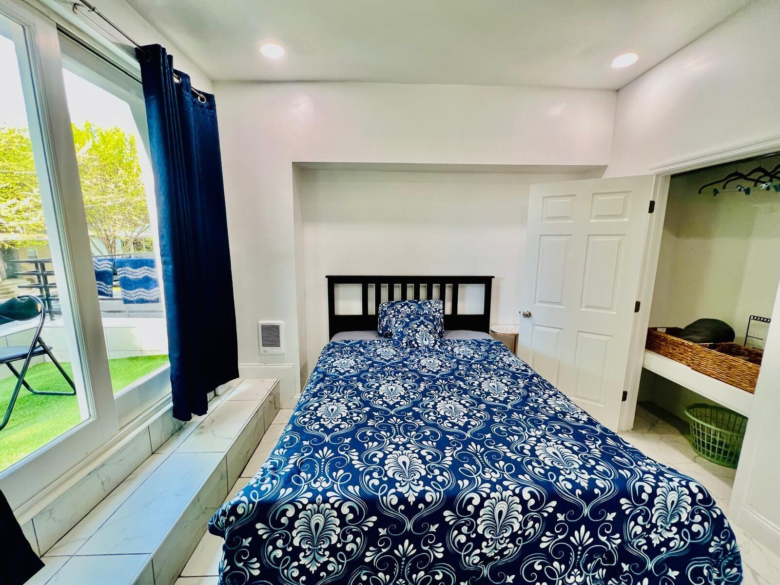 Un dormitorio con una cama grande, una ventana con cortinas, una puerta y una mesita con una cesta.