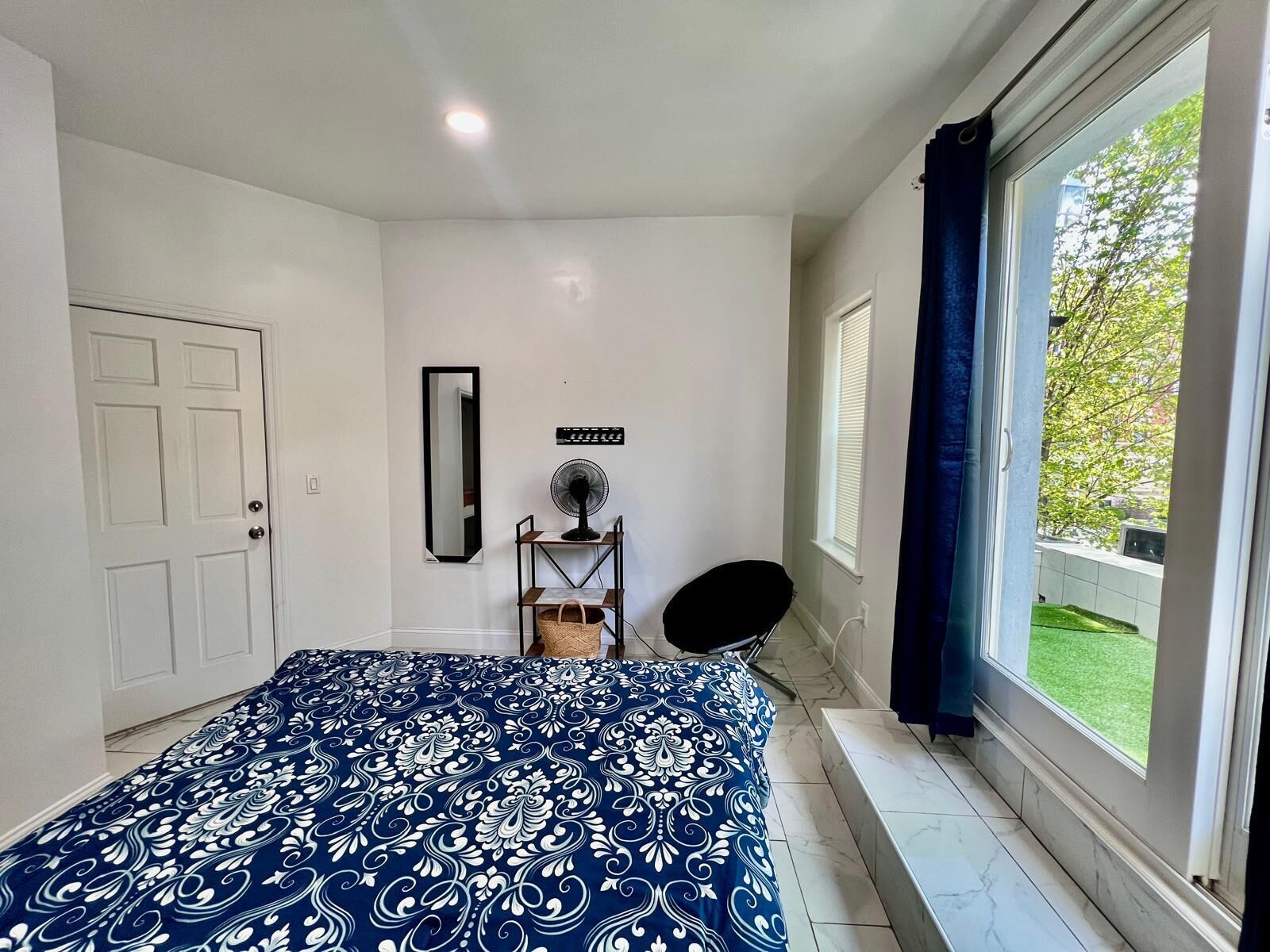 Ein Schlafzimmer mit einem großen Bett, einem Standventilator, einem Spiegel und einem Fenster mit Vorhängen.