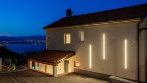A modern house with a sloped roof, a small balcony, and a view of a cityscape and water body at dusk.