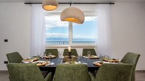 A dining room with a table set for six, green upholstered chairs, and a view of the sea through large windows.