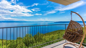 A balcony with a wicker chair, a metal railing, and a view of a large body of water and distant mountains.