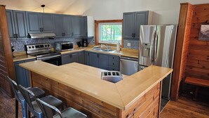 A modern kitchen with grey cabinets, stainless steel appliances, a wooden island with bar stools, and a view of the outdoors through a window.