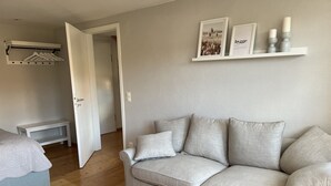Un soggiorno dai toni neutri con un divano color beige, un tavolino e un letto con copripiumino bianco. Una porta dà accesso a un'altra stanza.