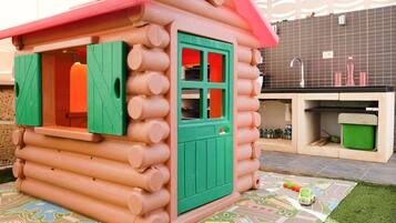 Una casita de madera con techo rojo y contraventanas verdes, un lavamanos y una alfombra de juego con diseño de carretera y vehículos.