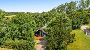 A small cabin with a deck, surrounded by dense trees and a gravel path.