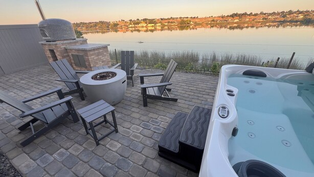 Un patio con mobiliario exterior, una bañera de hidromasaje y vistas a un lago con edificios al fondo.