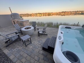 Un patio con mobiliario exterior, una bañera de hidromasaje y vistas a un lago con edificios al fondo.