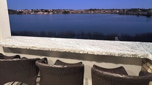 Un balcón con sillas de mimbre y un ventanal grande con vista a una masa de agua tranquila y una costa lejana.