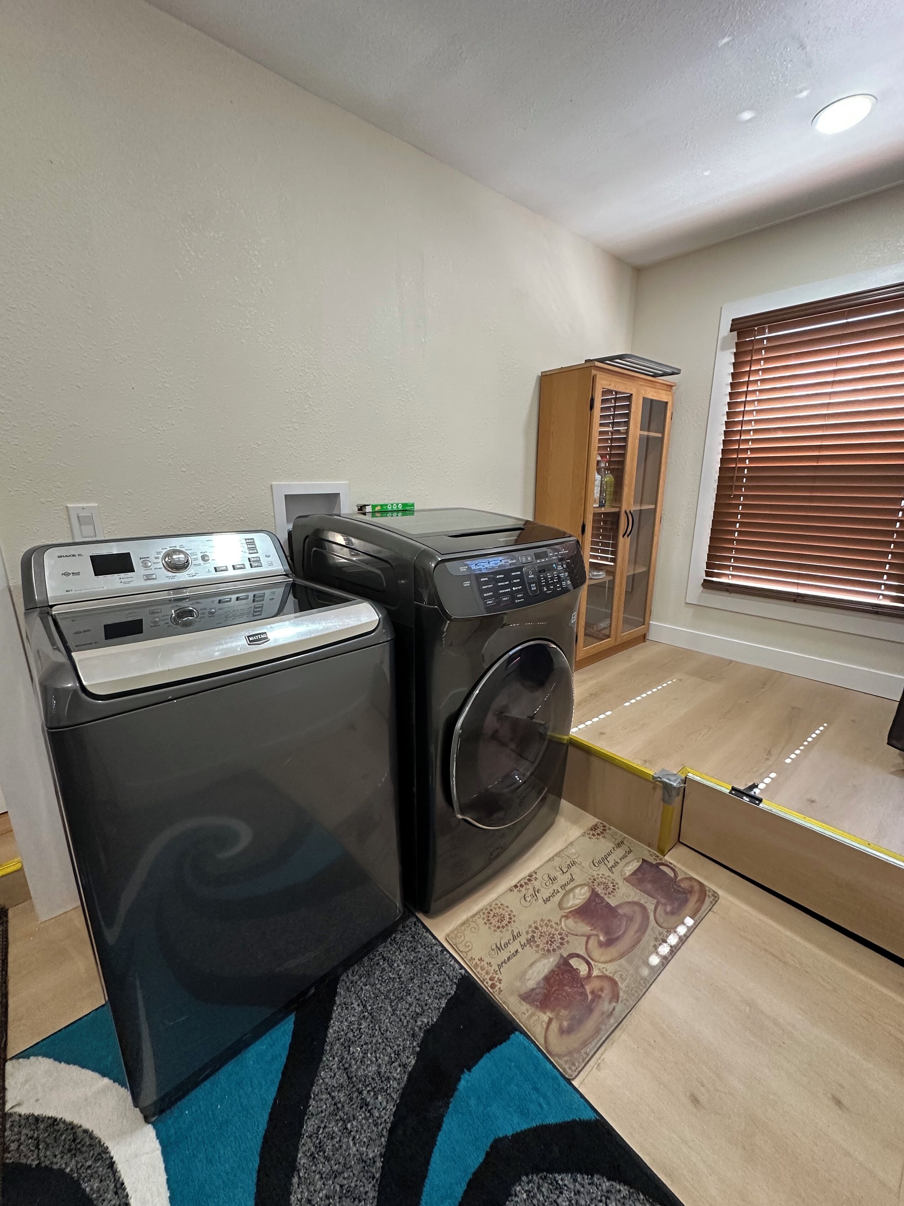 A laundry area with two washing machines, a wooden cabinet, and a window with blinds.