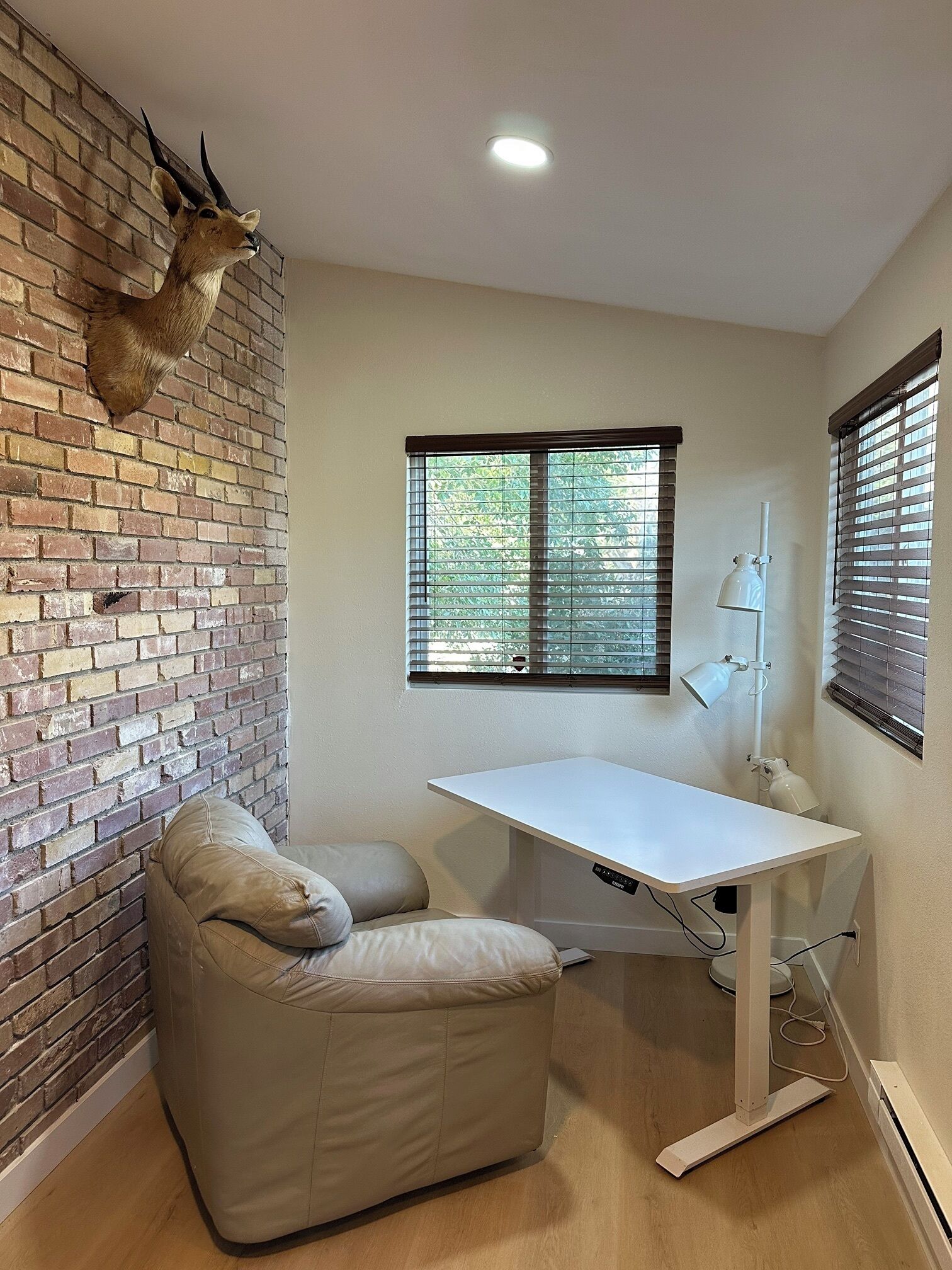 Une pièce avec un mur en briques, une tête d’animal empaillée accrochée au mur, un bureau avec une lampe, un fauteuil rembourré et une fenêtre avec des stores.
