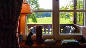 Ein Fenster mit Blick auf eine grüne Landschaft, eine Lampe, eine Bär-Figur und eine kleine Schachtel auf einer Holzoberfläche.