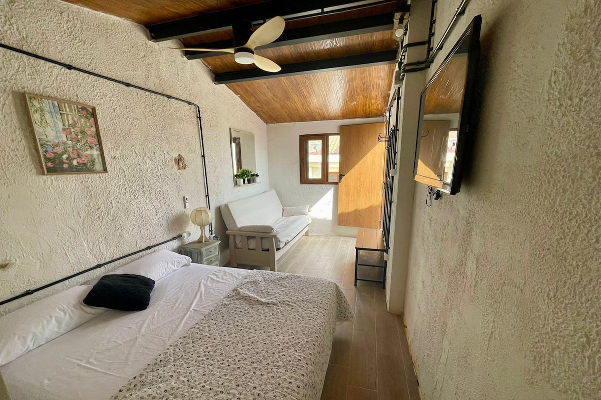 Ein schmales Zimmer mit einem Bett, einem kleinen Tisch und einem Stuhl. Eine Deckenlampe, ein Fenster und ein Spiegel sind an der Wand angebracht.