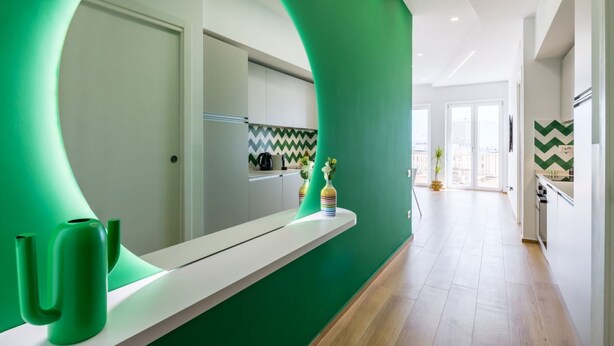 A modern kitchen with a green accent wall, a white countertop, and a mirror reflecting the space.
