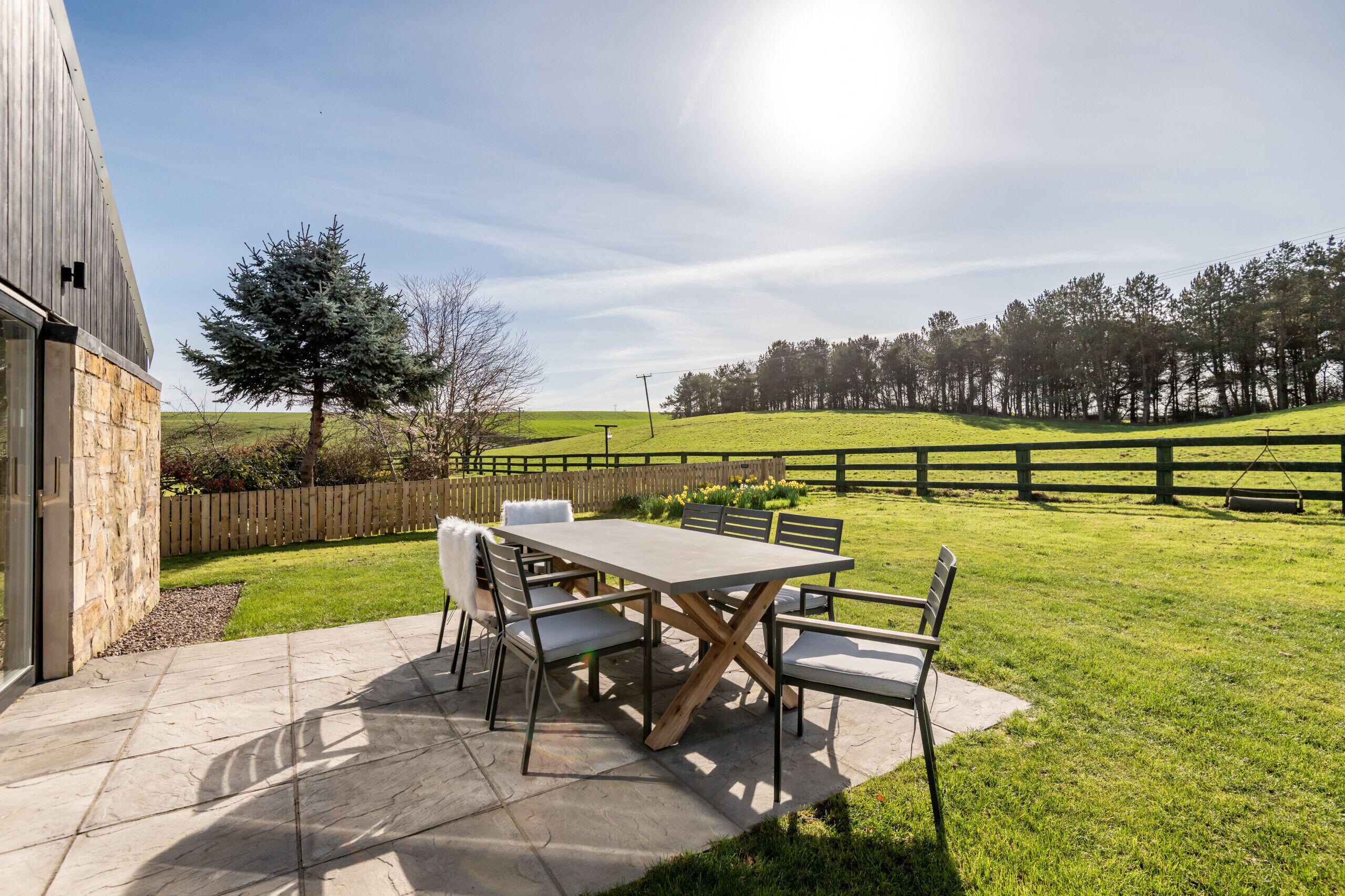 A patio with a table and chairs, a stone wall, and a view of a grassy field and trees.
