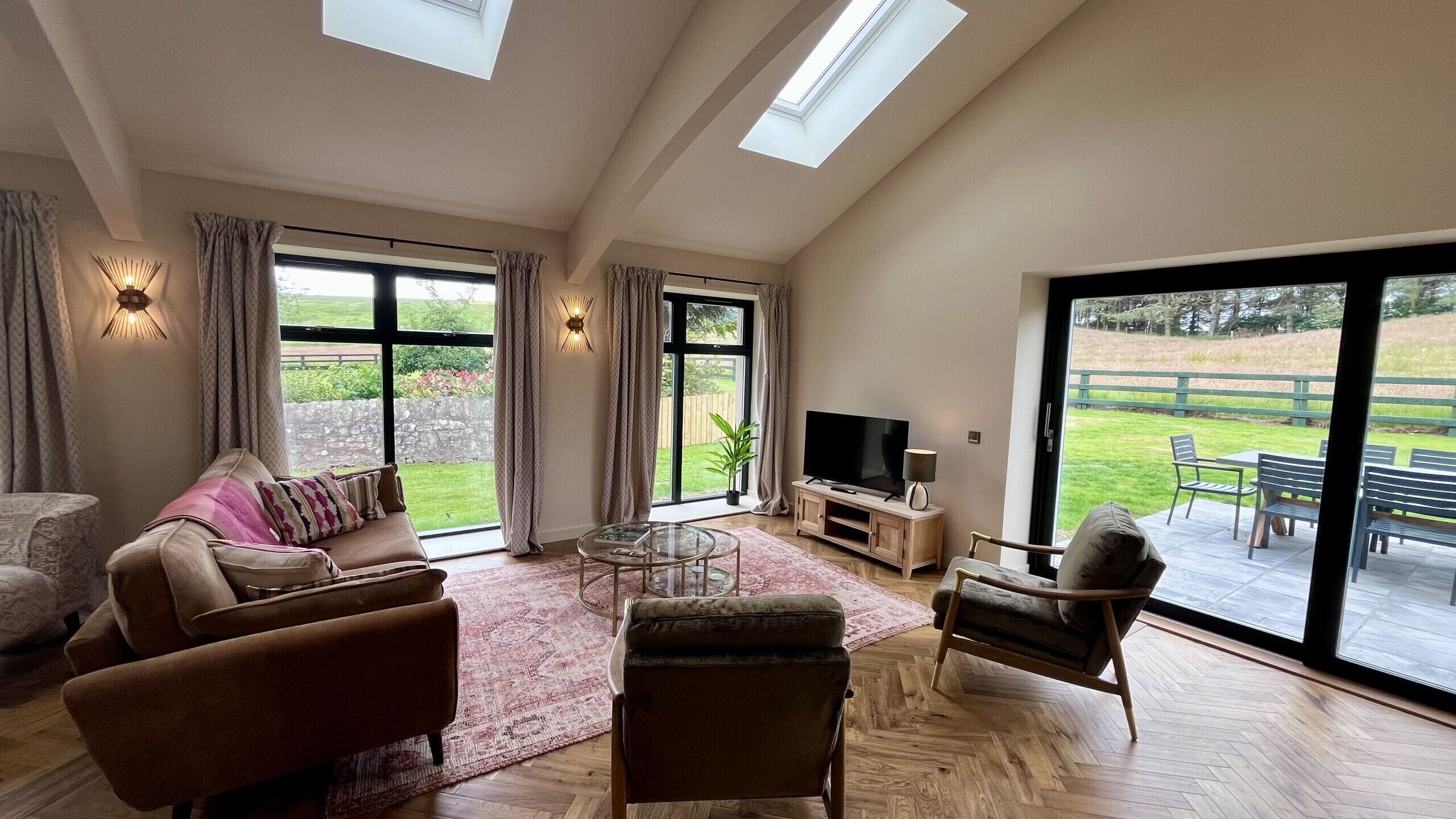 A spacious living room with a sofa, armchairs, a coffee table, and a TV. Large windows offer views of a garden and a wooden deck.