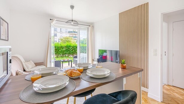 A modern dining area with a wooden table set for breakfast, a view of greenery outside, and a sliding door leading to a balcony. - Sweet cocoon at Disney (Montévrain)