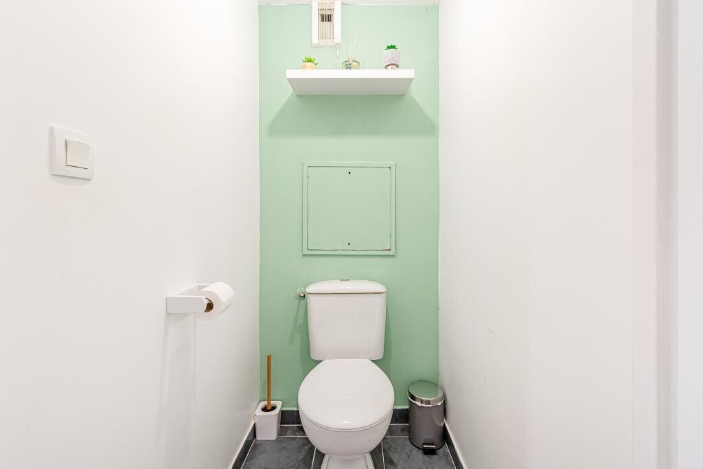 Et lille badeværelse med toilet, skraldespand, toiletpapirholder og en hylde med en plante.