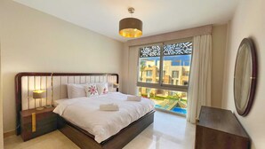 A modern hotel room with a large bed, a nightstand, a mirror, and a view of the city through a large window. - Xuru Stays I Homestyle 3BR Penthouse Pool & Beach View (El Gouna)