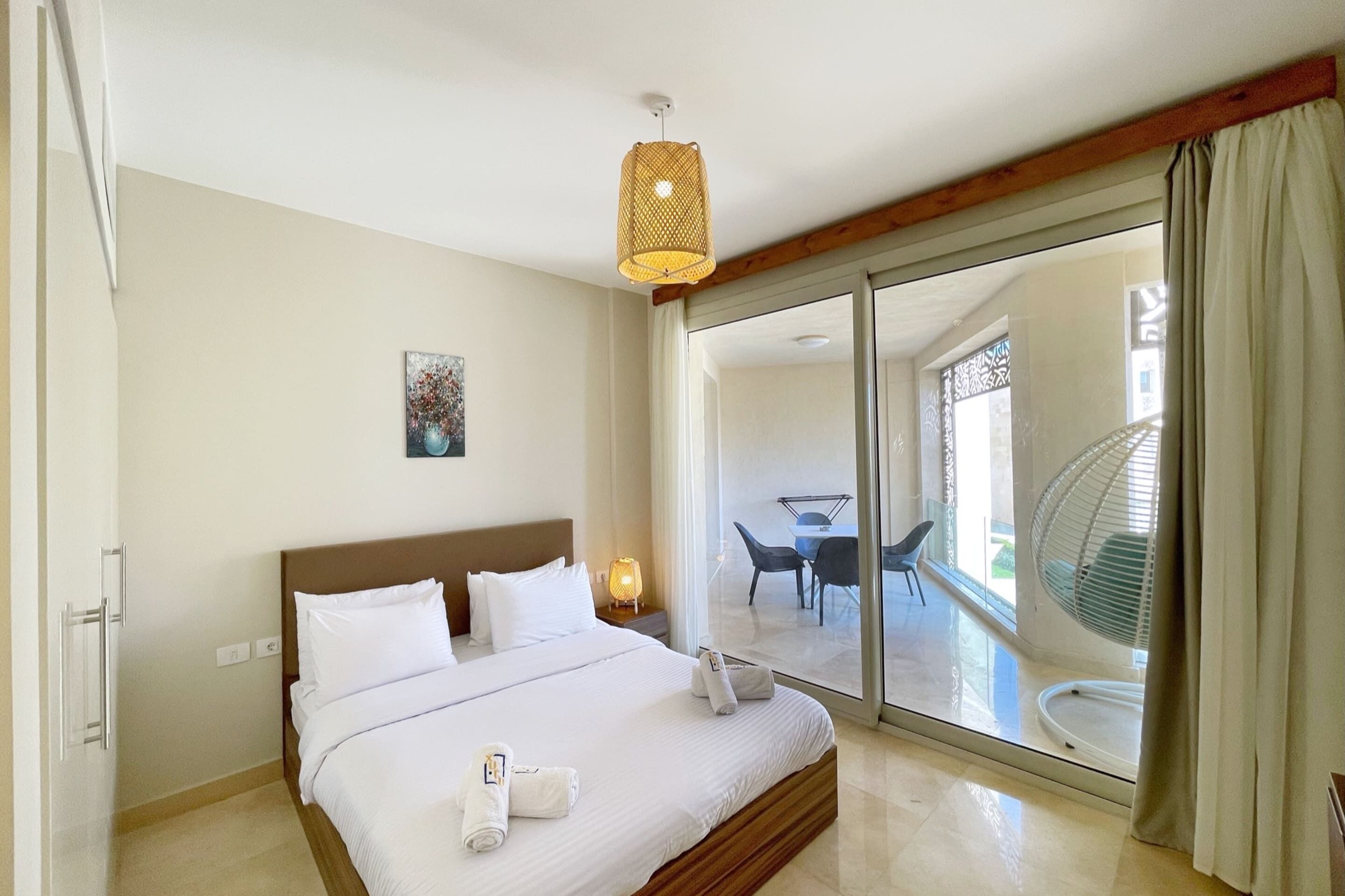 Habitación de hotel con una cama grande, una mesita de noche con lámpara, un cuadro en la pared y una puerta corrediza de vidrio que da a un balcón con mobiliario exterior.