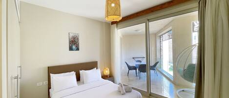 Habitación de hotel con una cama grande, una mesita de noche con lámpara, un cuadro en la pared y una puerta corrediza de vidrio que da a un balcón con mobiliario exterior.
