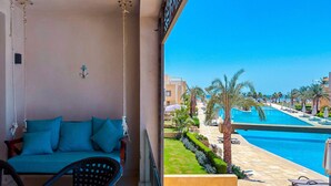 A balcony with a sofa and chairs, overlooking a swimming pool and palm trees. - Xuru Stays I Homestyle 3BR Penthouse Pool & Beach View (El Gouna)