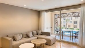 A modern living room with a beige sectional sofa, two round wooden tables, and a large sliding glass door leading to an outdoor pool area. - Xuru Stays I Homestyle 3BR Penthouse Pool & Beach View (El Gouna)