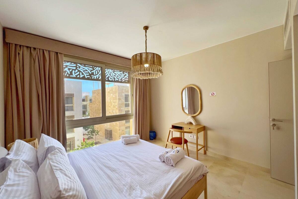 A hotel room with a bed, a desk, a mirror, and a view of buildings outside the window.
