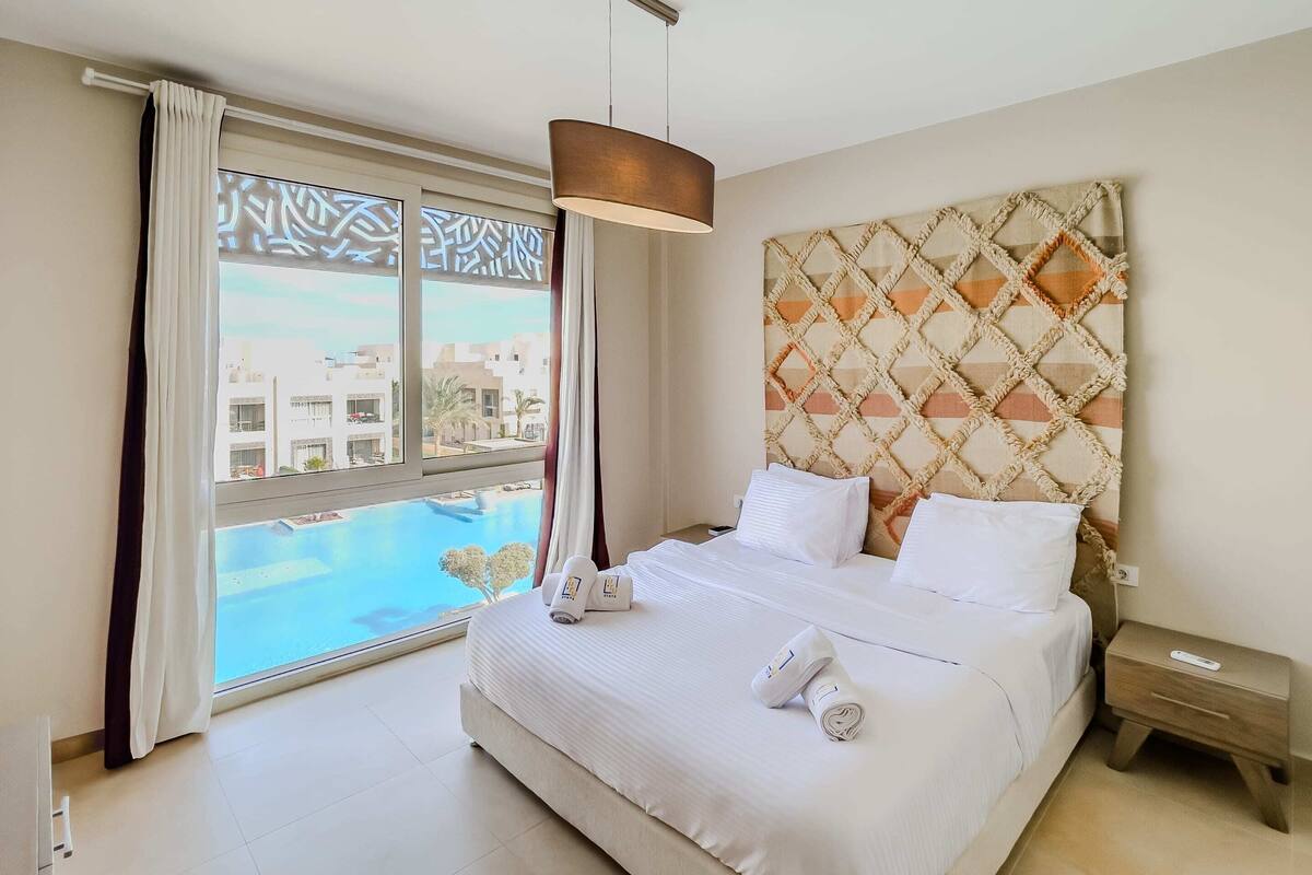 A hotel room with a large bed, a headboard with a geometric pattern, a bedside table, and a view of a swimming pool through a large window.