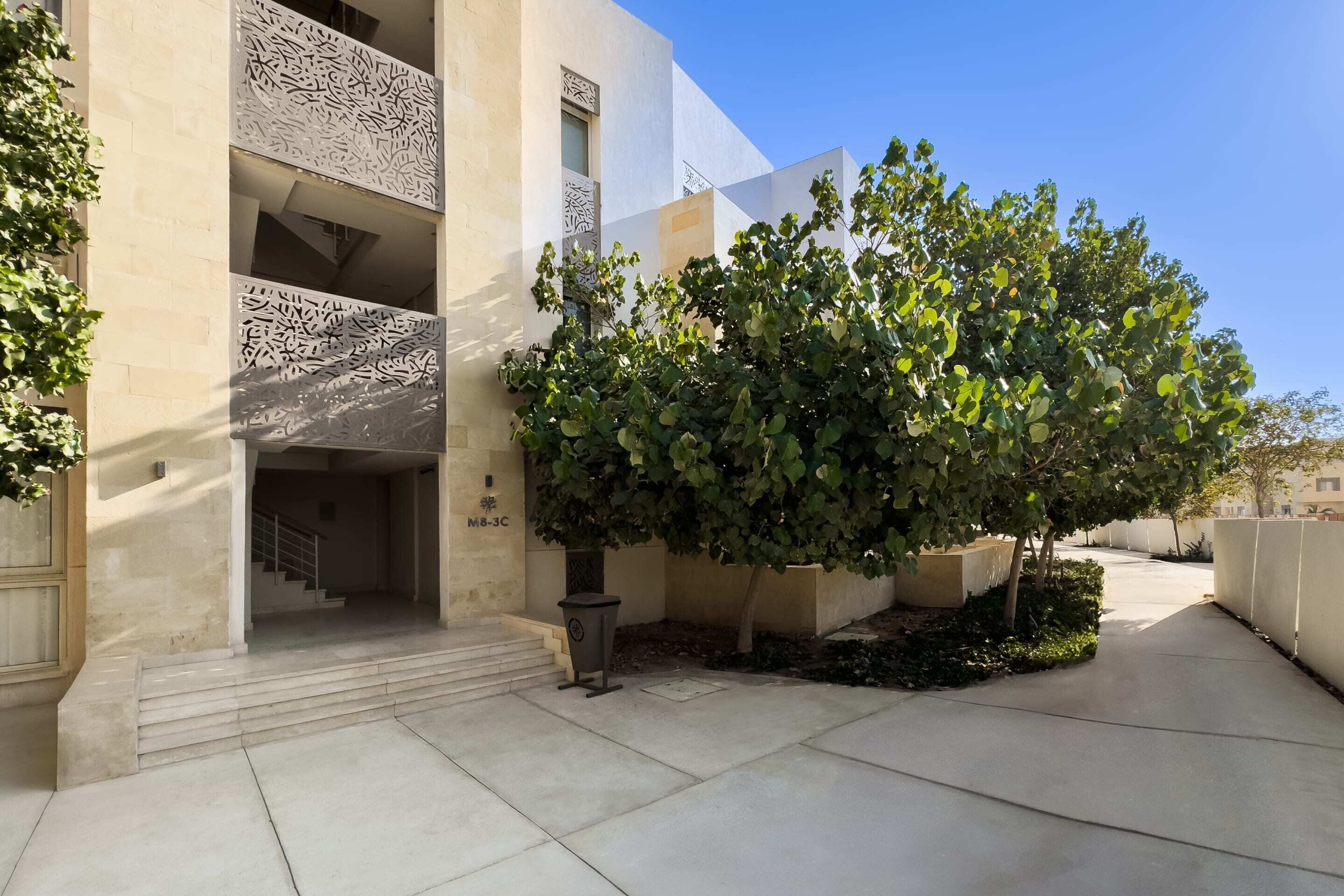 Un edificio con un diseño arquitectónico moderno, que presenta una escalera hacia una entrada, un árbol en el patio y un contenedor de basura.