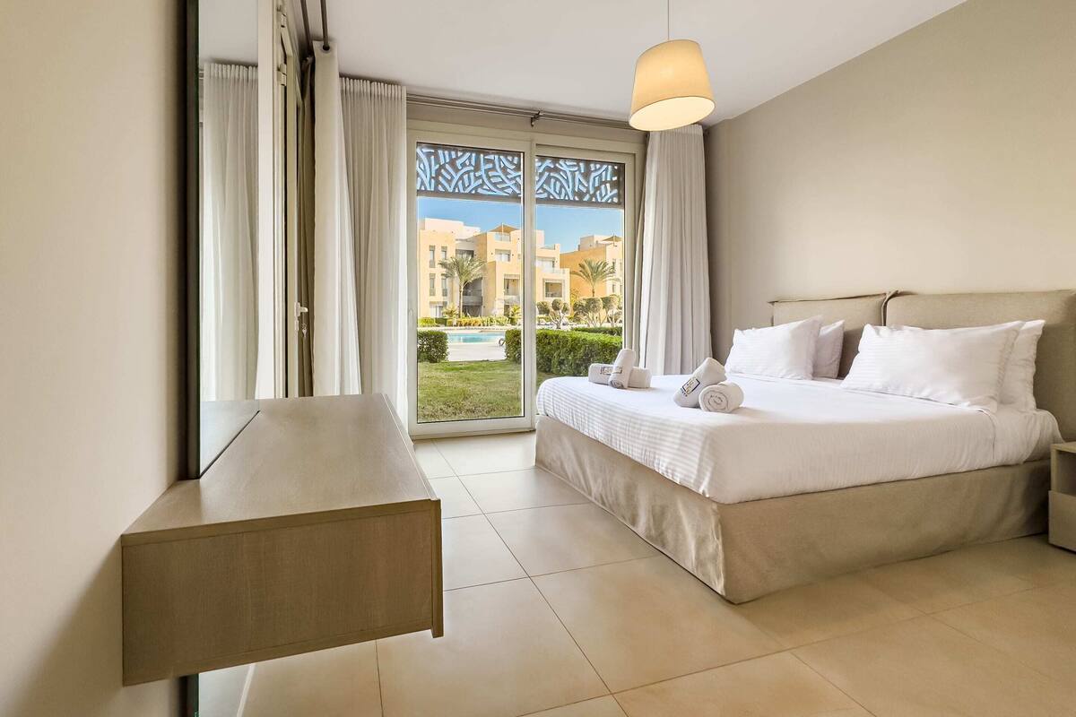 A hotel room with a bed, bedside table, and a view of a pool and buildings.