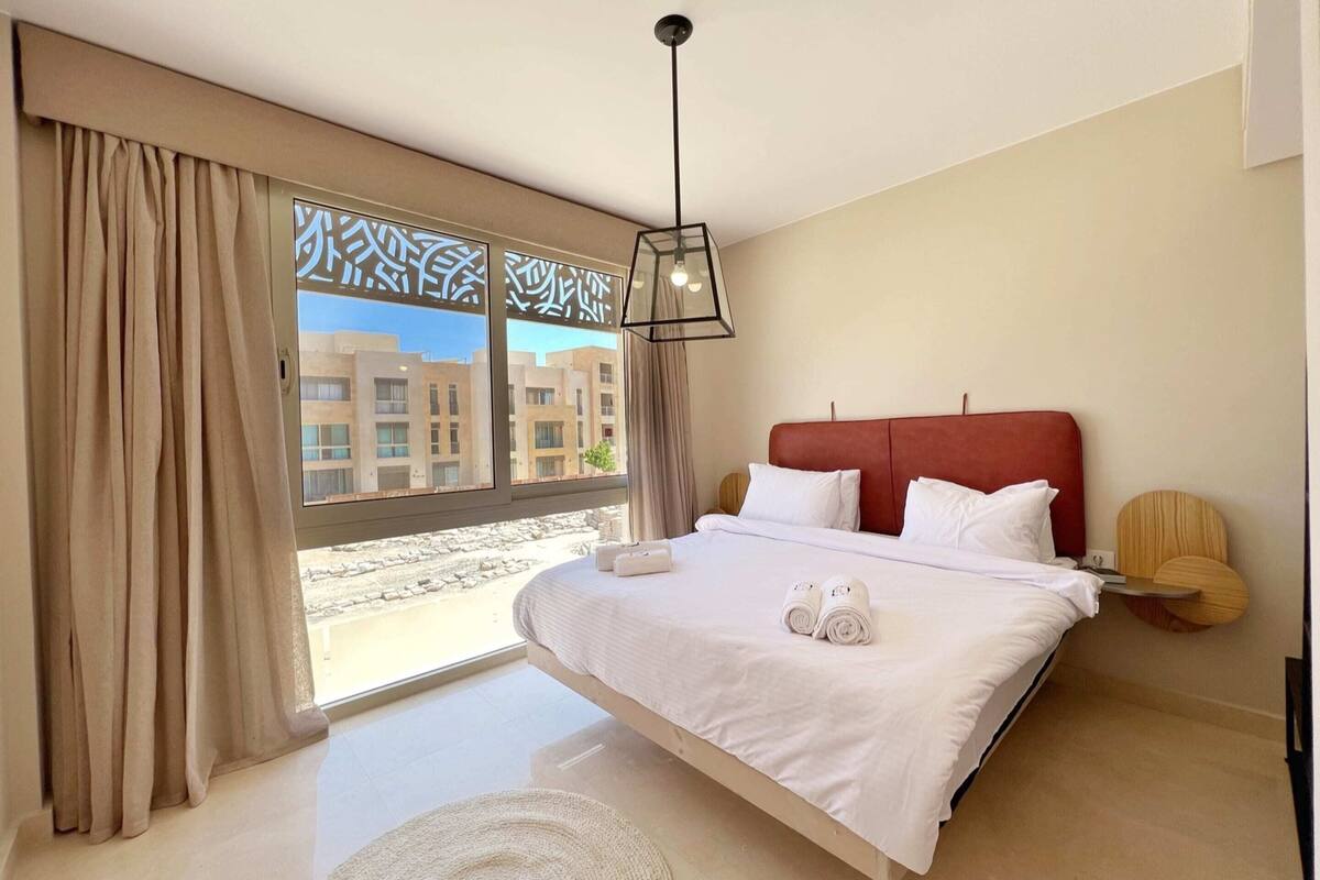 A modern hotel room with a large bed, a red headboard, and a view of buildings through large windows.