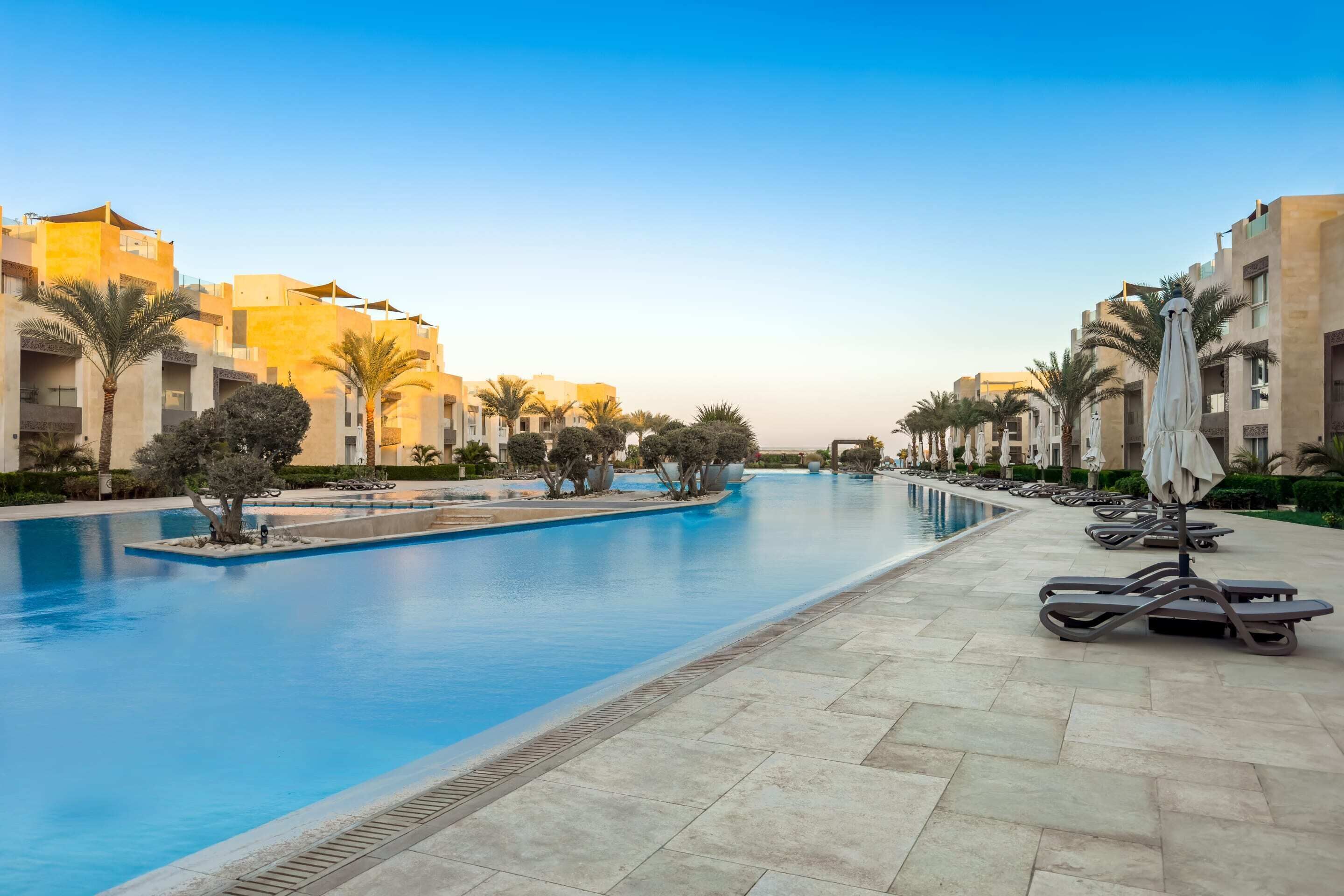 A swimming pool with lounge chairs and umbrellas, surrounded by palm trees and modern buildings.