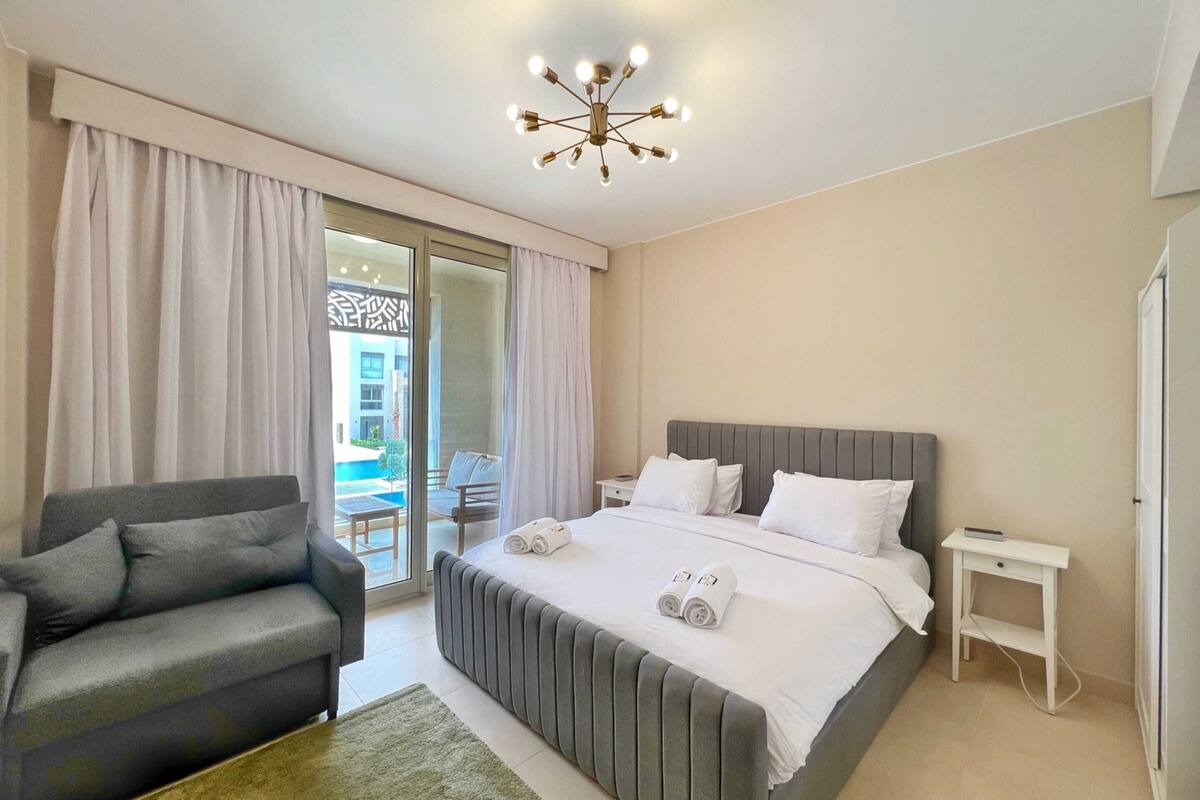 A modern hotel room with a large bed, a grey sofa, a small bedside table, and a view of an outdoor pool.