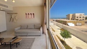 A modern living room with a sofa, a coffee table, and a view of the cityscape through large windows. - Xuru Stays I Homestyle & Summery 1bdr@ Mangroovy Free Beach Pool (El Gouna)