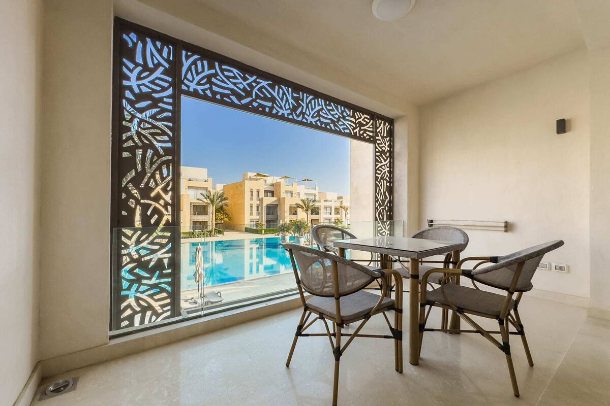 A room with a large window overlooking a swimming pool and buildings. There is a table with chairs and a small decorative screen.