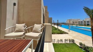 A balcony with two white cushioned chairs and a wooden table, overlooking a swimming pool and a well-maintained lawn. - Xuru Stays I Homestyle & Summery 1bdr@ Mangroovy Free Beach Pool (El Gouna)